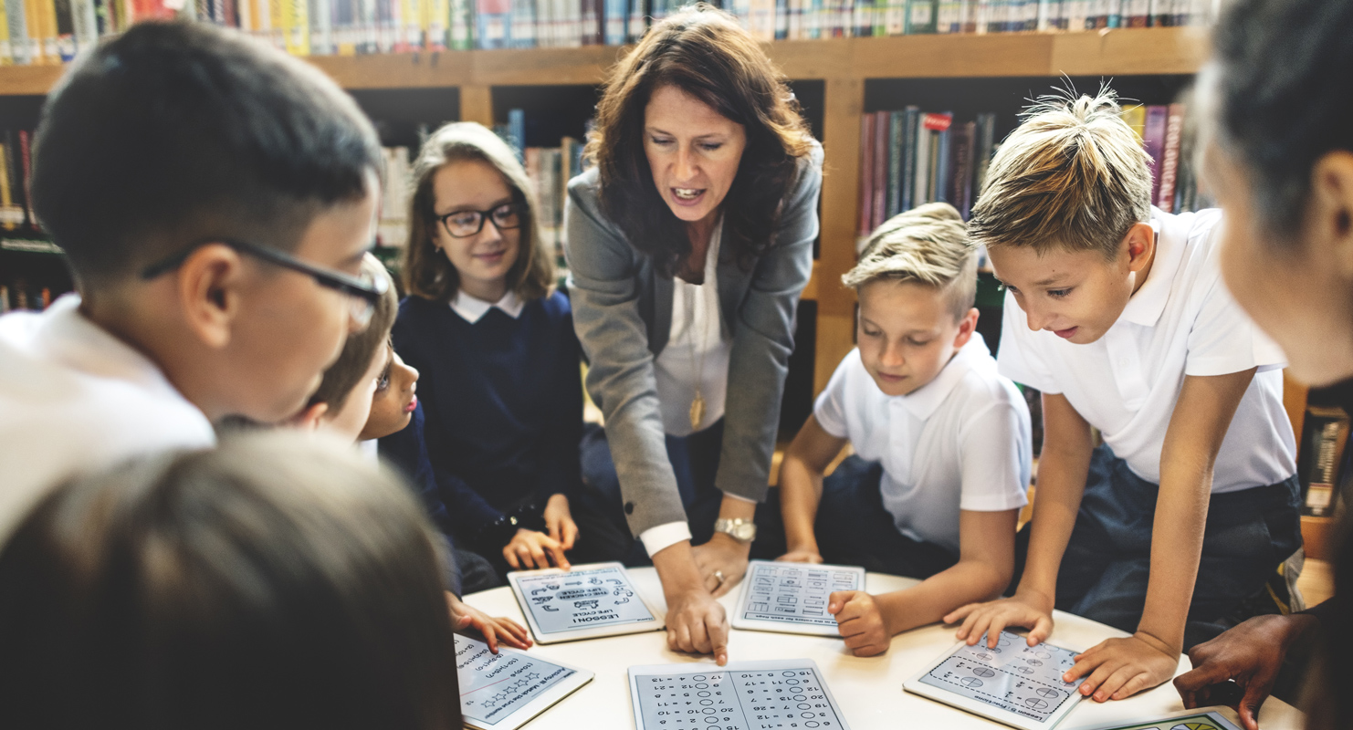 A teacher and a group of 6 students, each with a tablet, discuss something across a round table.