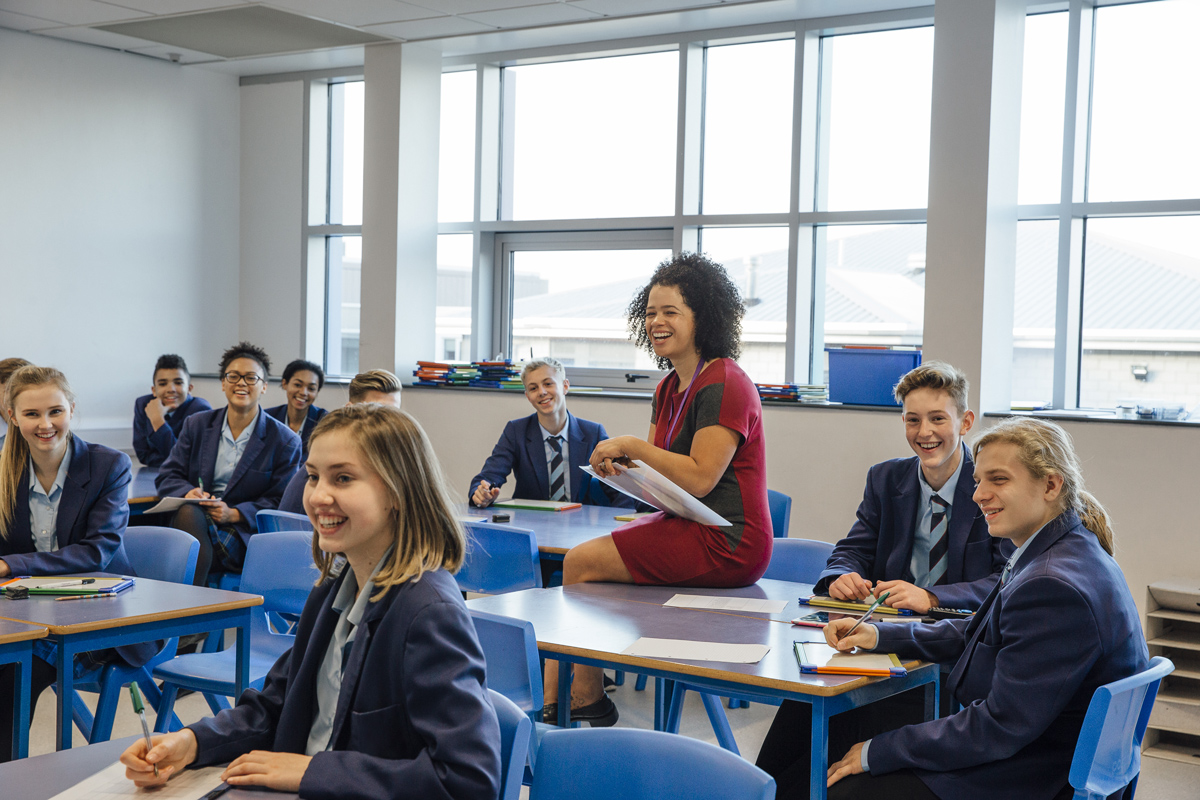 A classroom of secondary school students laughing, their teacher is sitting on a students desk laughing too